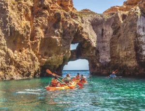 Grupo em passeio de kayak pelas grutas e formações rochosas da costa algarvia, navegando em águas cristalinas azul-turquesa junto às falésias douradas do Algarve.