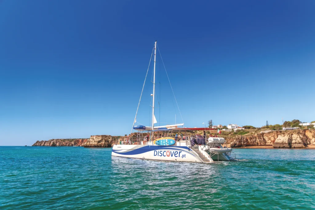 Catamarã da Discover Tours a navegar ao longo da costa algarvia, com falésias douradas, mar calmo e céu azul intenso no Algarve.